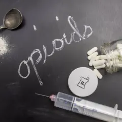 Napis "opioidy" na tablicy, obok łyżeczki z proszkiem, strzykawki, kapsułek i symbolu recepty.