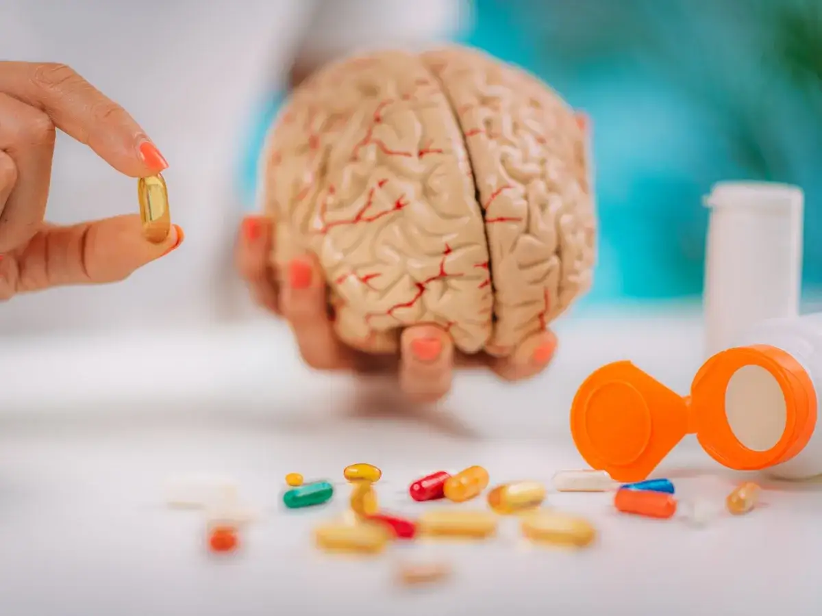 Dłoń trzyma kapsułkę obok modelu mózgu, wokół rozsypane tabletki. Czy to początek podróży w świat psychodelików?