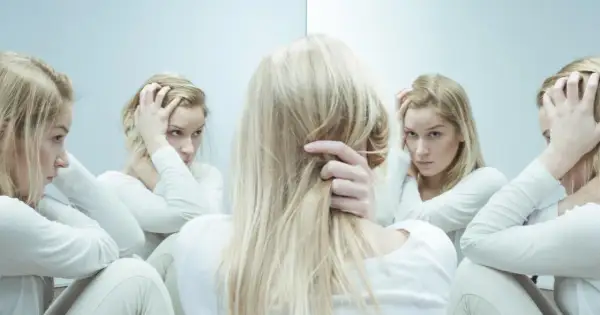 Kobieta w białej bluzie z długimi blond włosami, zmagająca się z kwetaplex, widzi swoje odbicia w lustrze, każde z innym wyrazem twarzy.