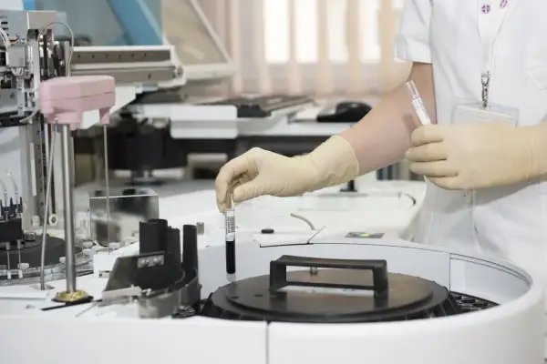 Ręka w rękawiczce umieszcza probówkę z ciemnym płynem w aparacie laboratoryjnym. To co to za badanie?